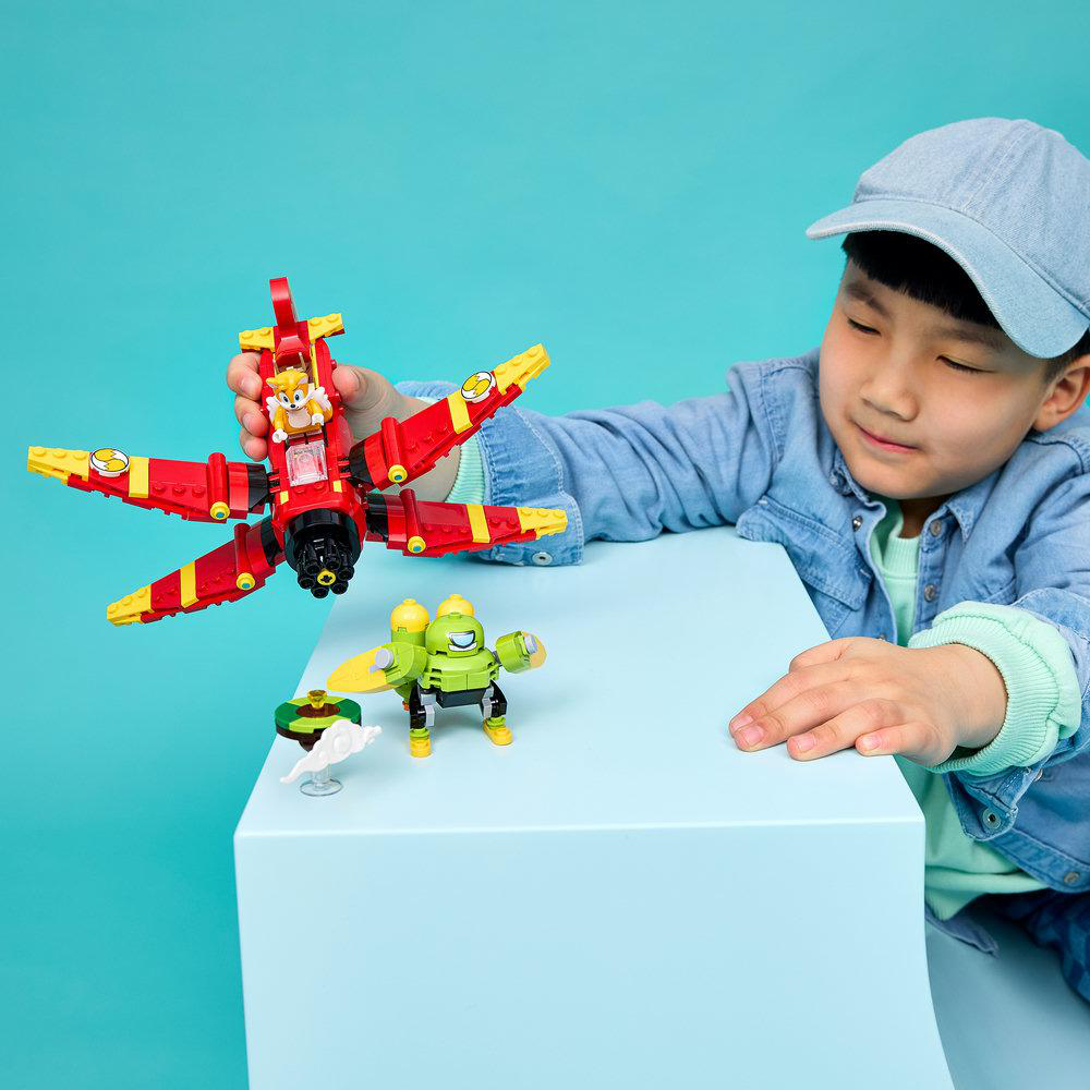 Ein Junge spielt mit einem rot-gelben Lego-Flugzeug und einem grünen Roboterspielzeug auf einem blauen Tisch.