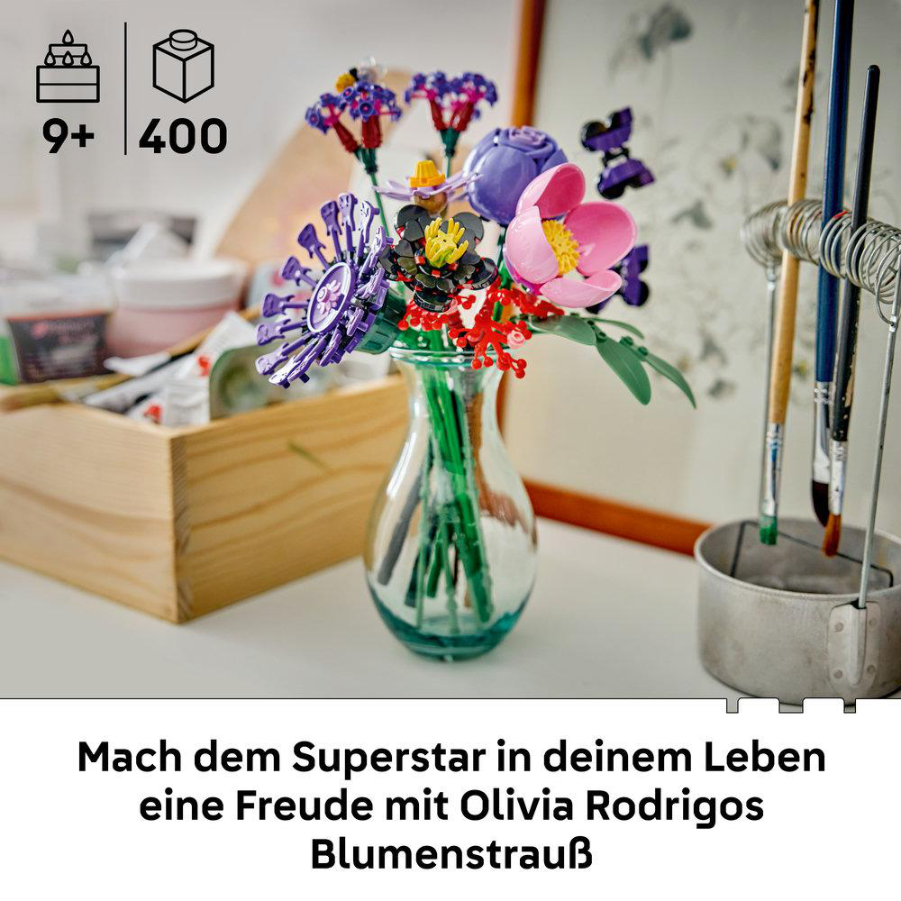 Ein bunter LEGO Blumenstrauß in einer Glasvase auf einem Tisch mit Kunstbedarf im Hintergrund.
