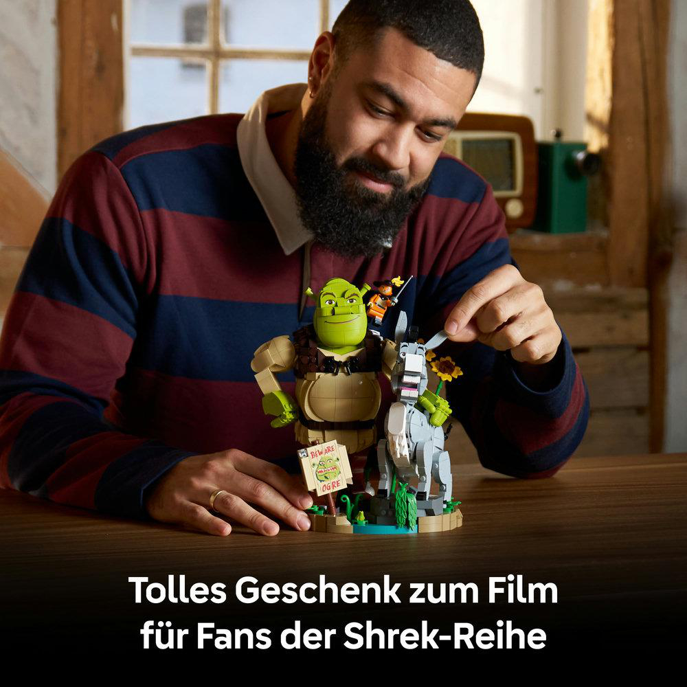 Ein Mann baut ein Shrek-LEGO-Set mit Shrek, Esel und Stiefelkater an einem Holztisch zusammen.