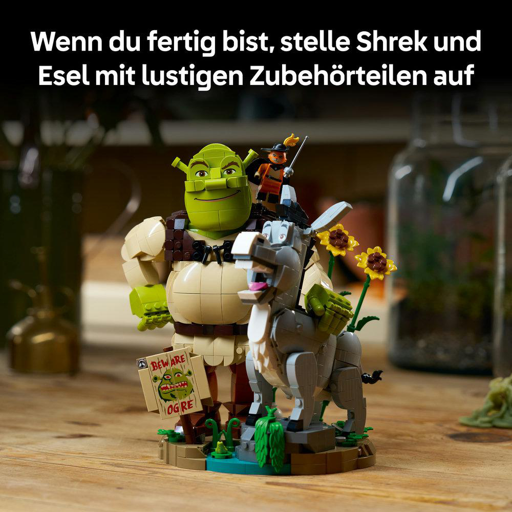 Lego-Figuren von Shrek, Esel und dem gestiefelten Kater auf einem hölzernen Tisch.