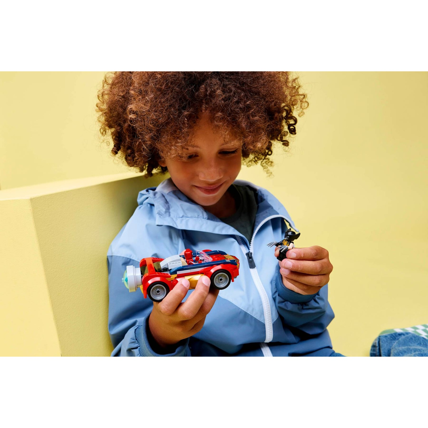 Un bambino riccio gioca con un'auto giocattolo Lego e un piccolo action figure su sfondo giallo.