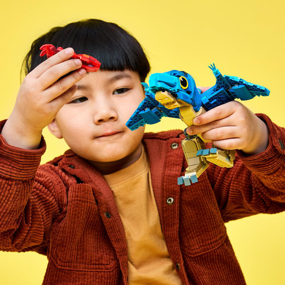 Ein Kind in einem braunen Hemd spielt vor gelbem Hintergrund mit roten und blauen Spielzeugdinosauriern.