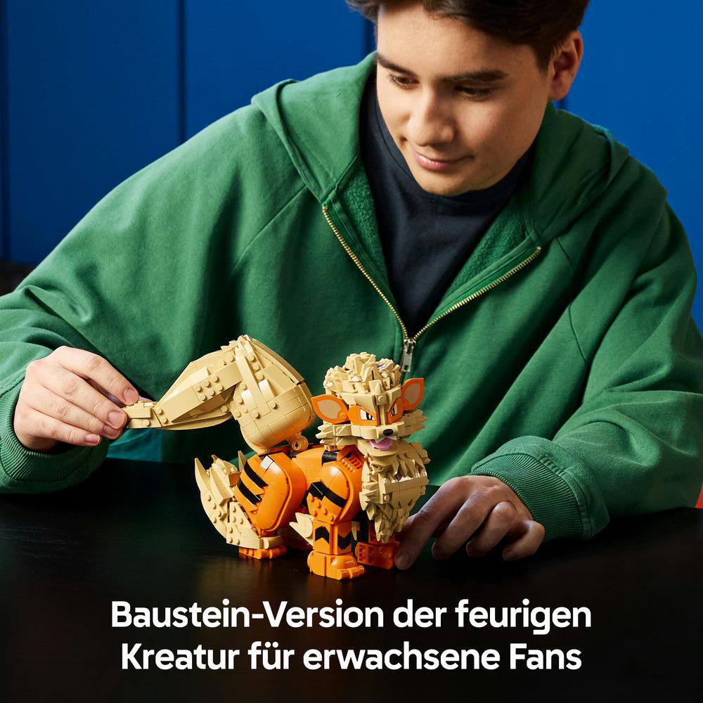 Ein junger Mann im grünen Hoodie baut eine orange und beige Lego-Kreatur auf einem schwarzen Tisch vor blauem Hintergrund.