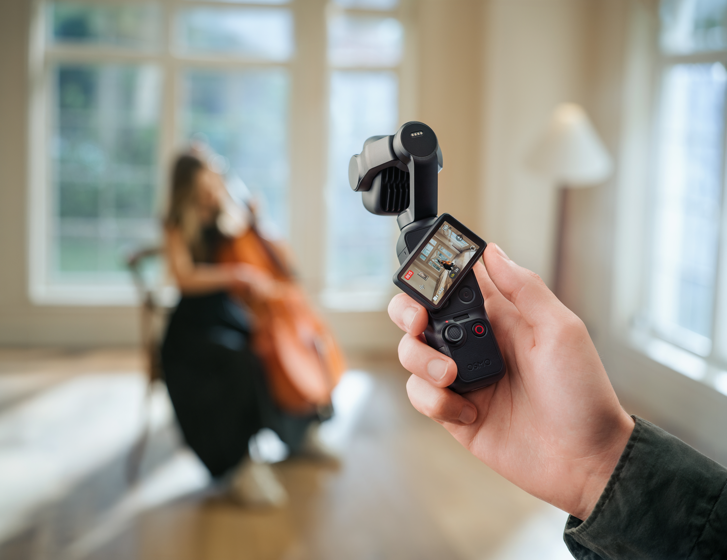Una mano sostiene una cámara Osmo Pocket negra, con una mujer tocando el violonchelo al fondo.