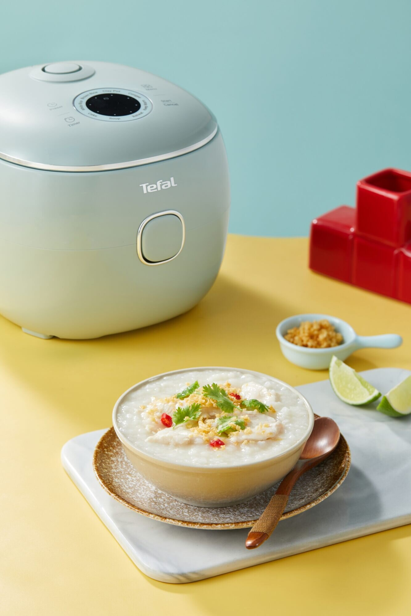 Jasnozielony ryżowar Tefal obok miski congee przystrojonej ziołami, kawałkami mięsa i chili.