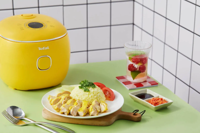 Żółty ryżowar Tefal, hainanczyński kurczak z ryżem na talerzu, napój i sosy na stole.