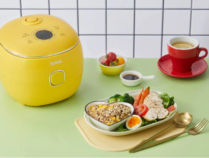 Żółty ryżowar Tefal obok posiłku z kaszą, kurczakiem i warzywami oraz czerwona filiżanka.