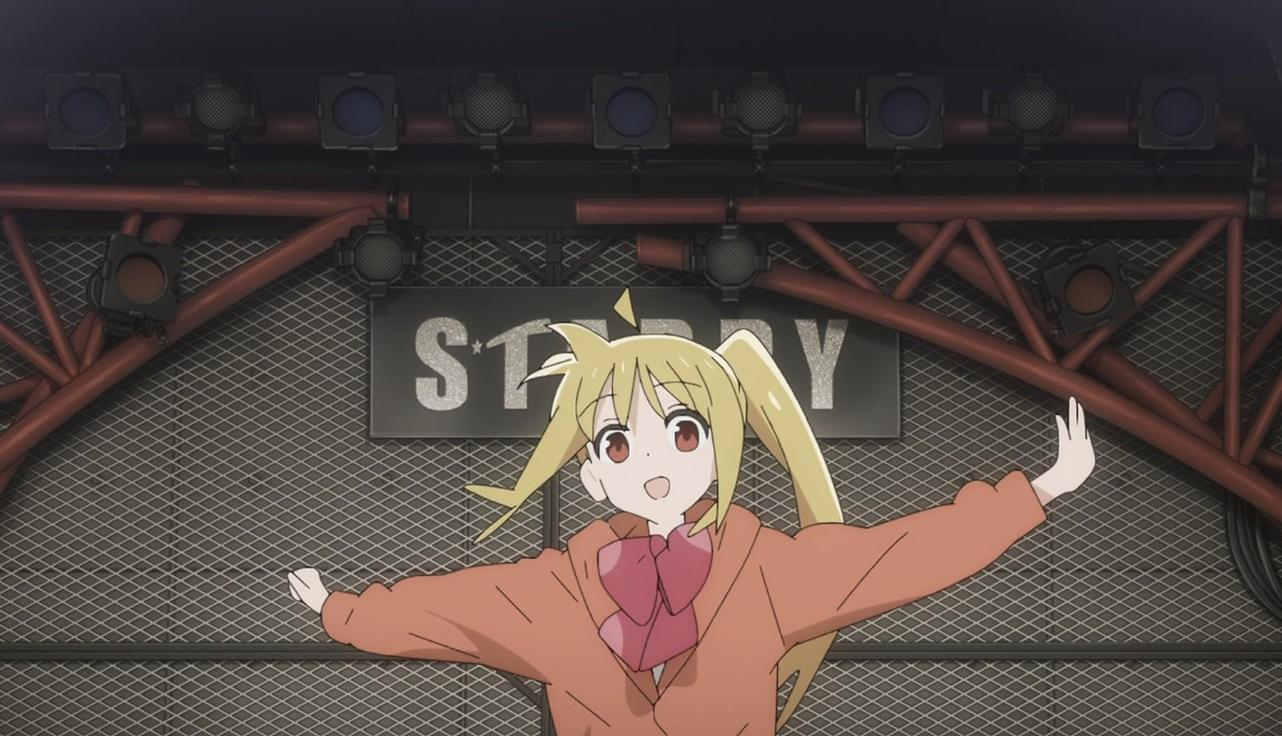 Ein blondes Anime-Mädchen mit Zöpfen und roter Schleife steht freudig vor einer Bühne mit Scheinwerfern.