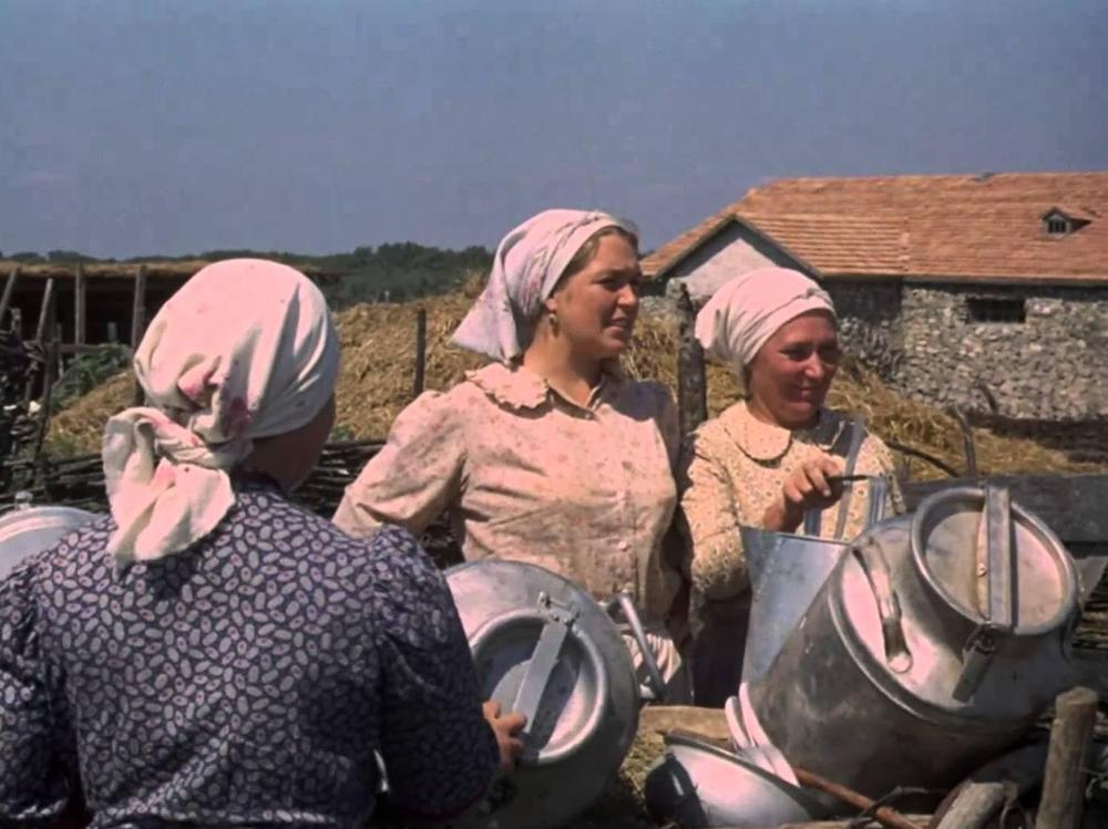 Drei Frauen in Kopftüchern und Arbeitskleidung stehen mit großen Milchannen auf einem ländlichen Hof vor Gebäuden.