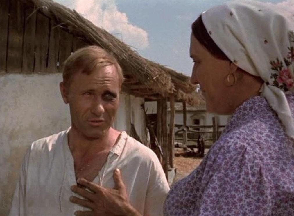Ein Mann mit blauem Auge spricht mit einer Frau in Tracht vor einer Hütte.
