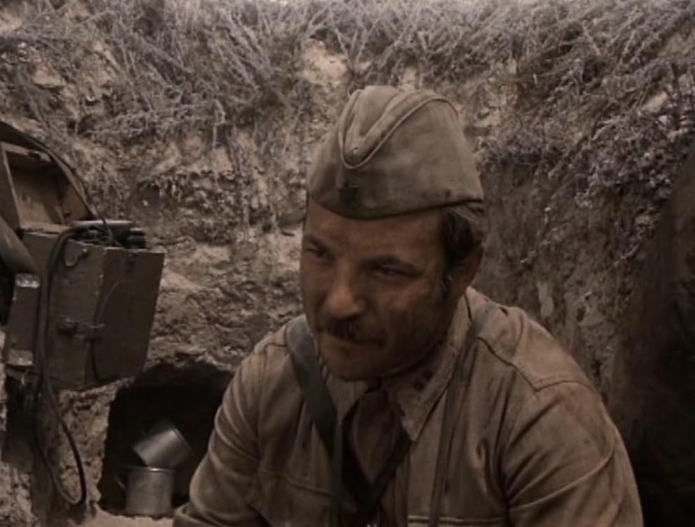 Ein Soldat in historischer Uniform und Feldmütze sitzt in einem Erdbunker mit Ausrüstung im Hintergrund.