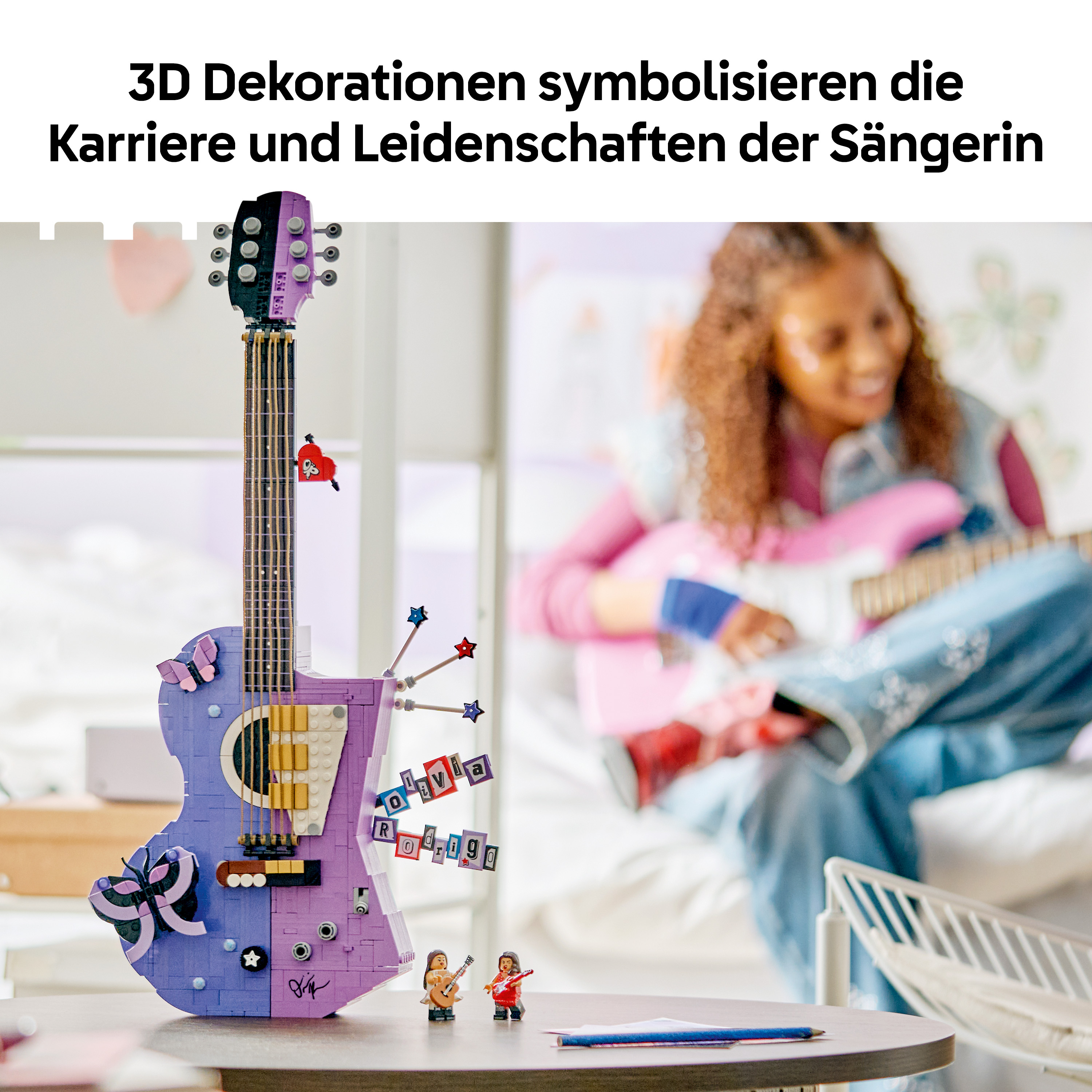 Eine gebaute Lego-Gitarre in Lila und Blau steht auf einem Tisch, im Hintergrund spielt ein Mädchen Gitarre.