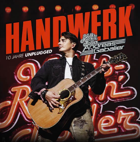 Andreas Gabalier hält eine Akustikgitarre vor einem neonbeleuchteten Hintergrund mit rotem Schriftzug Handwerk.