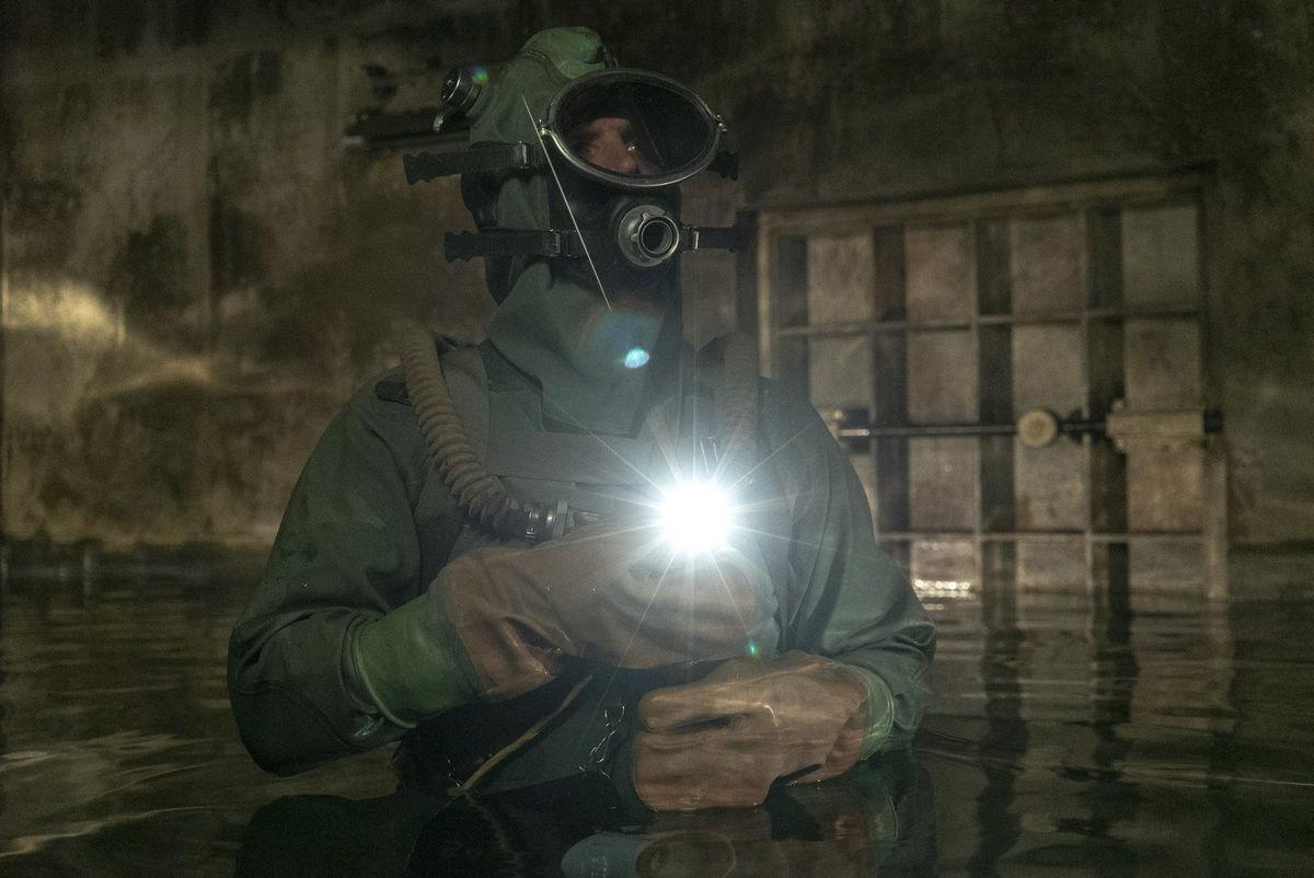 Ein Taucher in schwerem Schutzanzug und Gasmaske steht in dunkel gefluteten Tunneln und hält eine helle Taschenlampe.