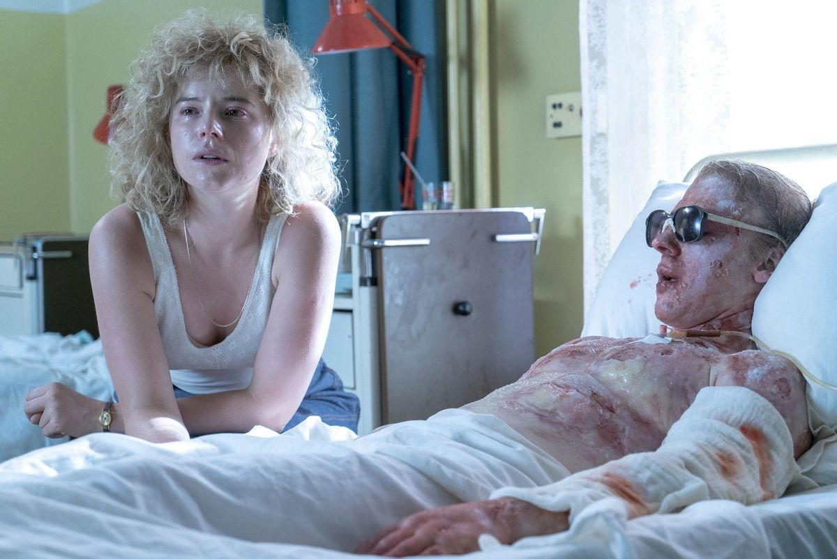 Eine blonde Frau sitzt besorgt am Krankenbett eines schwer verletzten Mannes mit Sonnenbrille.