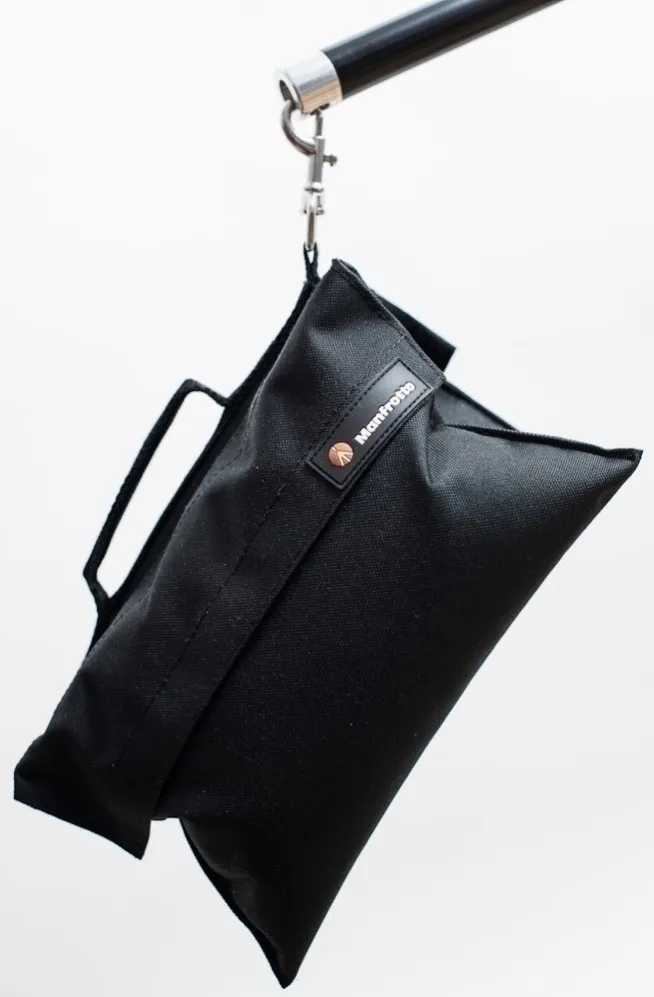 Un sac de sable noir Manfrotto suspendu à un crochet argenté sur fond blanc.