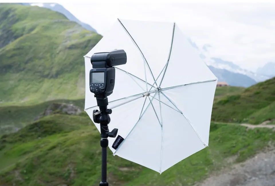 Un flash con un ombrello riflettente bianco su un treppiede davanti a un paesaggio montano.
