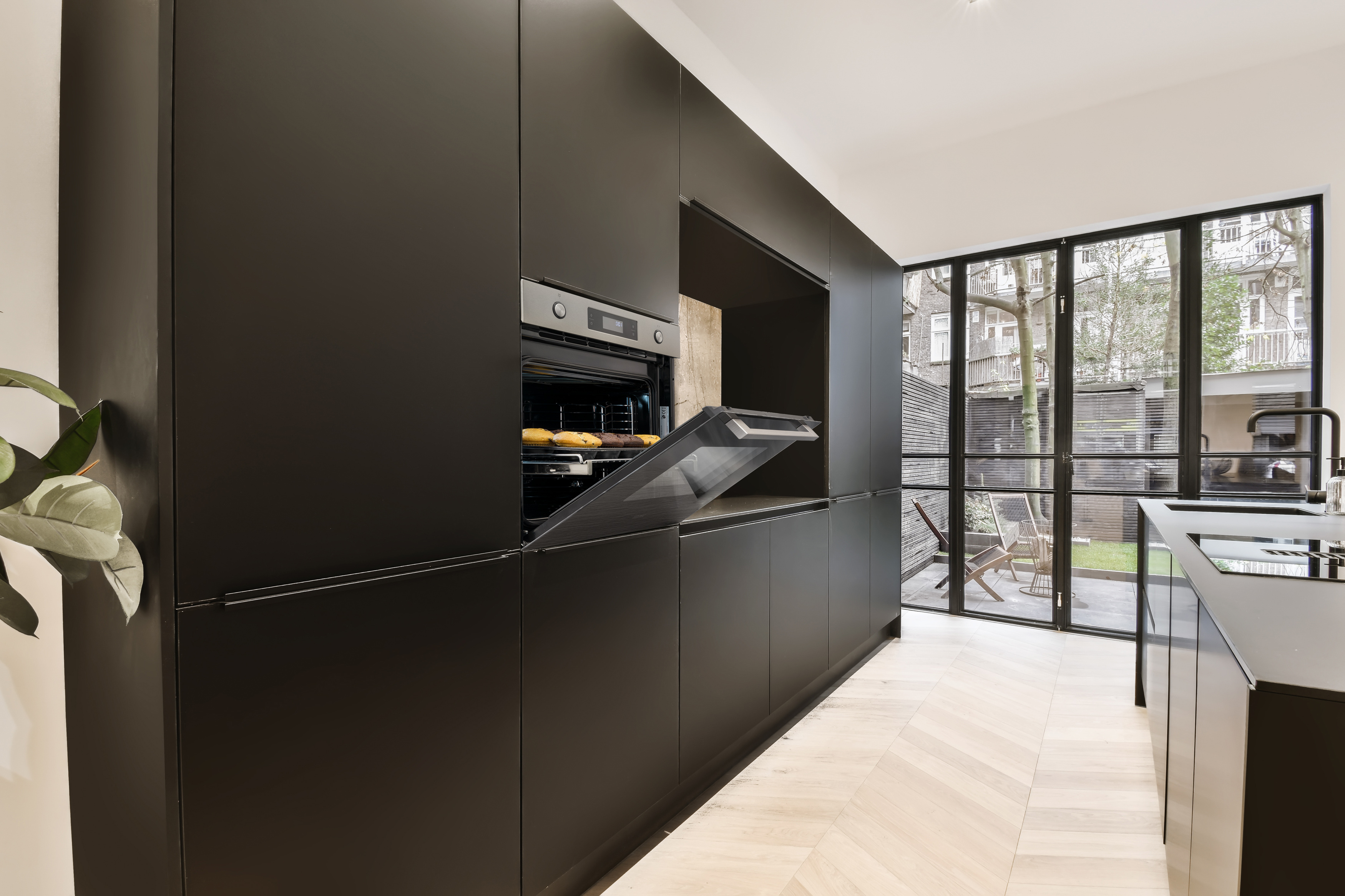 Moderne schwarze Küche mit eingebautem Ofen und Blick auf den Garten durch eine Glastür.