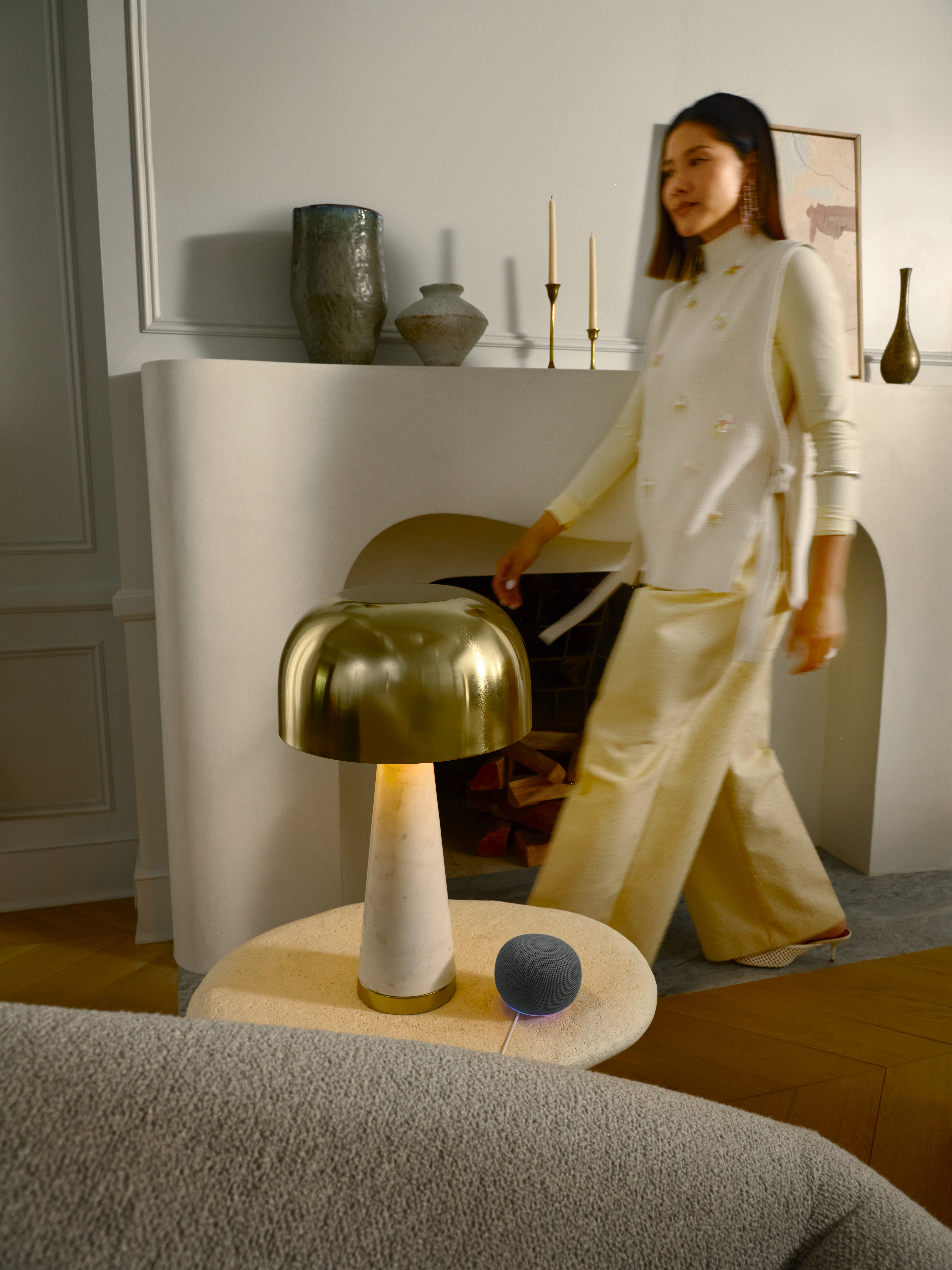 Eine Frau in gelber Kleidung geht im Hintergrund, während eine goldene Lampe auf einem Tisch im Vordergrund steht.