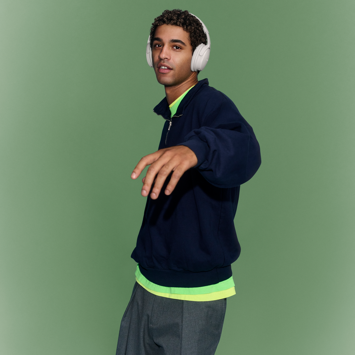Un jeune homme portant un casque audio a une veste bleu foncé, un t-shirt vert néon et un pantalon gris sur fond vert.
