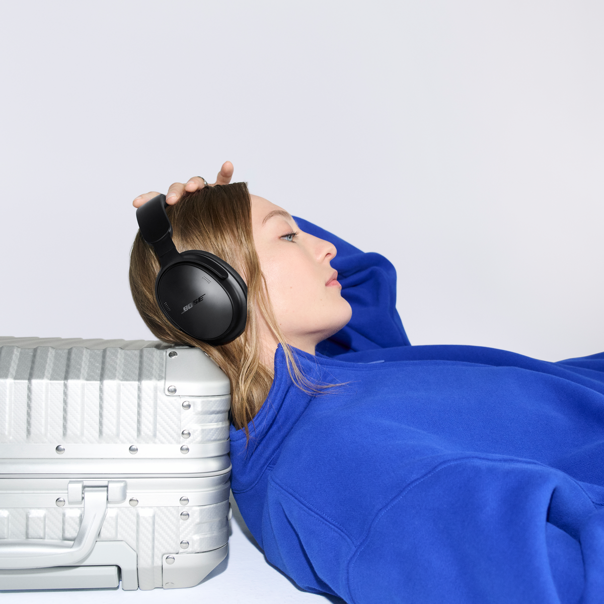 Une femme en pull bleu portant un casque noir pose sa tête sur une valise argentée contre un mur blanc.