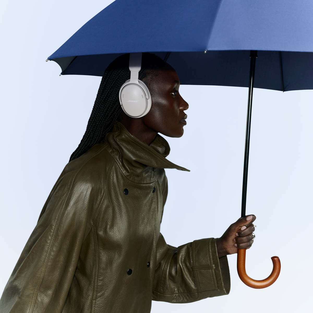 Une femme en manteau vert avec un casque blanc tient un parapluie bleu sur fond clair.