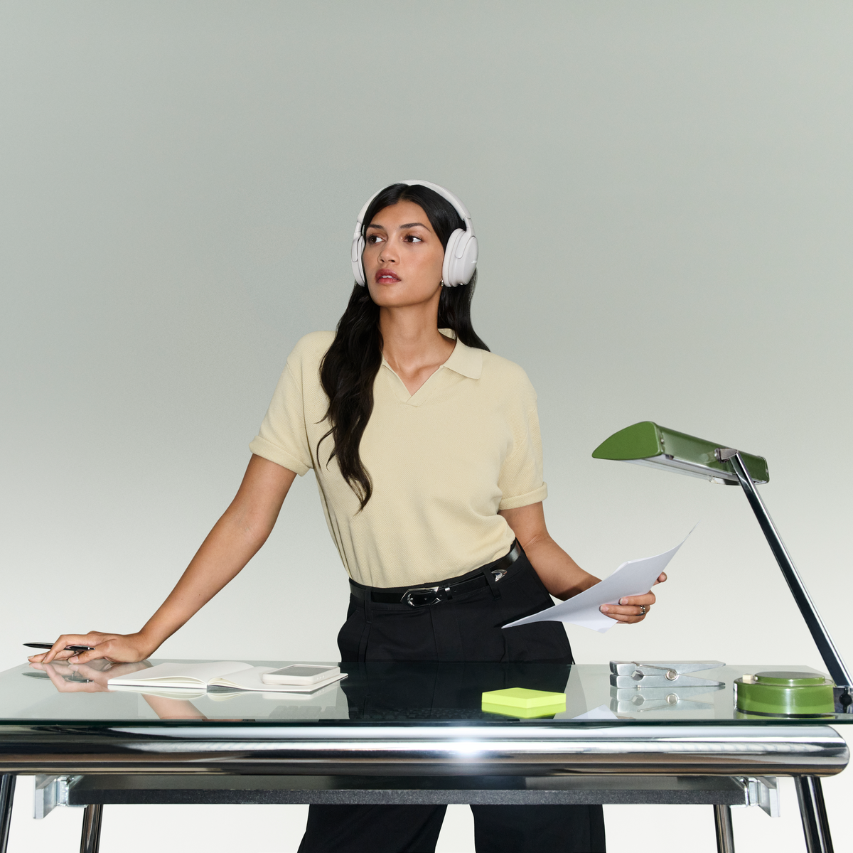 Une femme avec un casque est debout à un bureau en verre avec une lampe et des notes, regardant sur le côté.