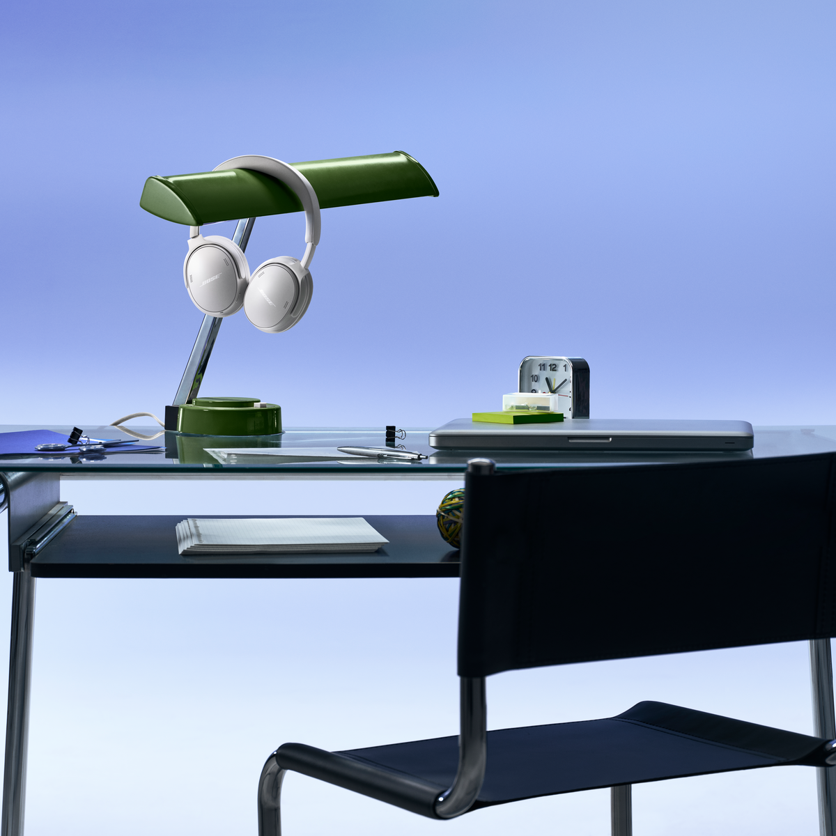 Un bureau en verre avec lampe verte, casque blanc, ordinateur portable et chaise sur fond bleu.
