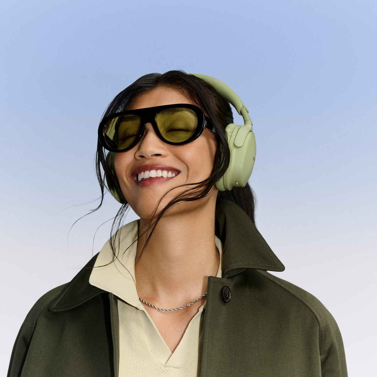 Une femme souriante aux cheveux foncés porte un casque vert, des lunettes de soleil et un manteau vert olive.