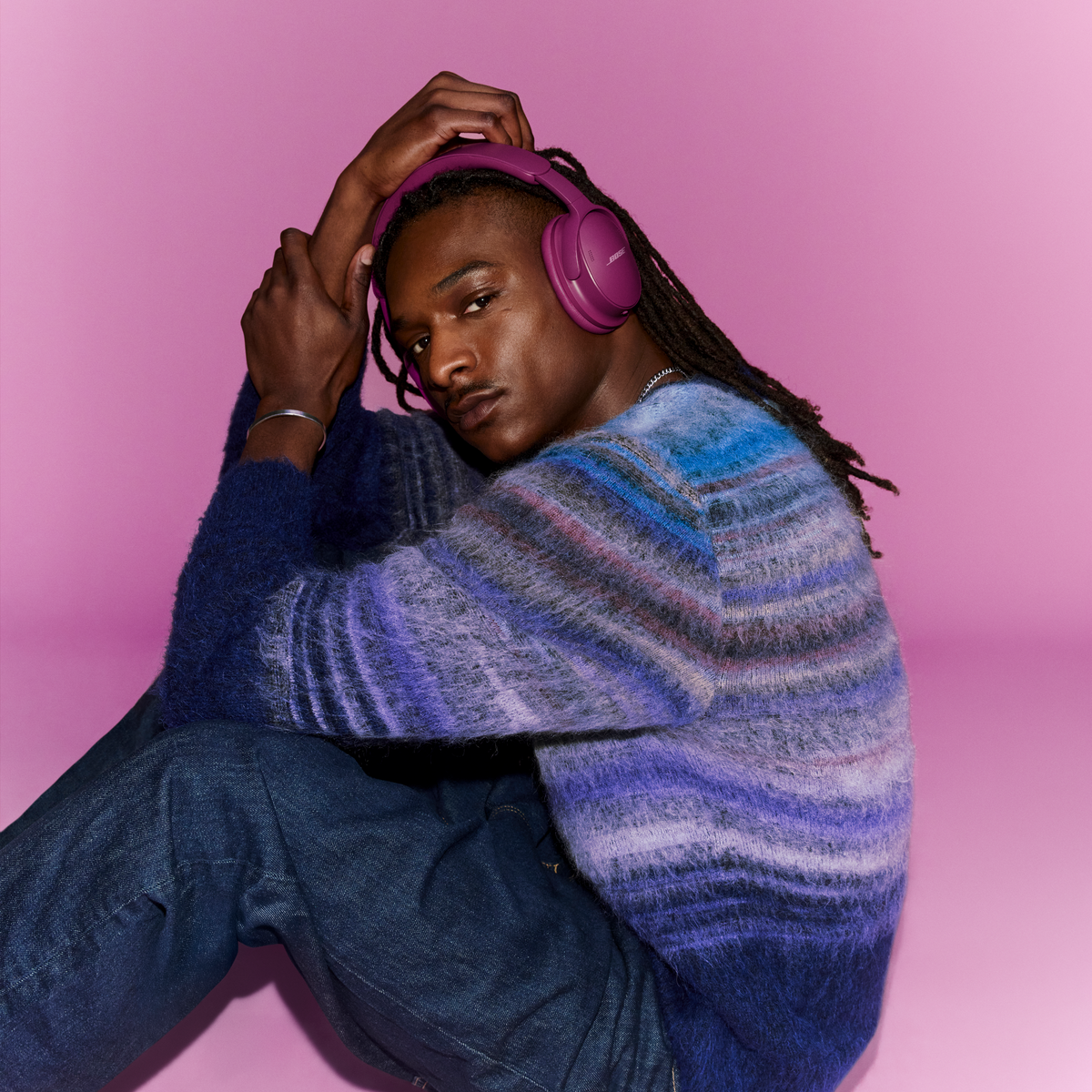 Un homme avec des dreadlocks et un casque violet porte un pull à rayures bleues sur fond rose.