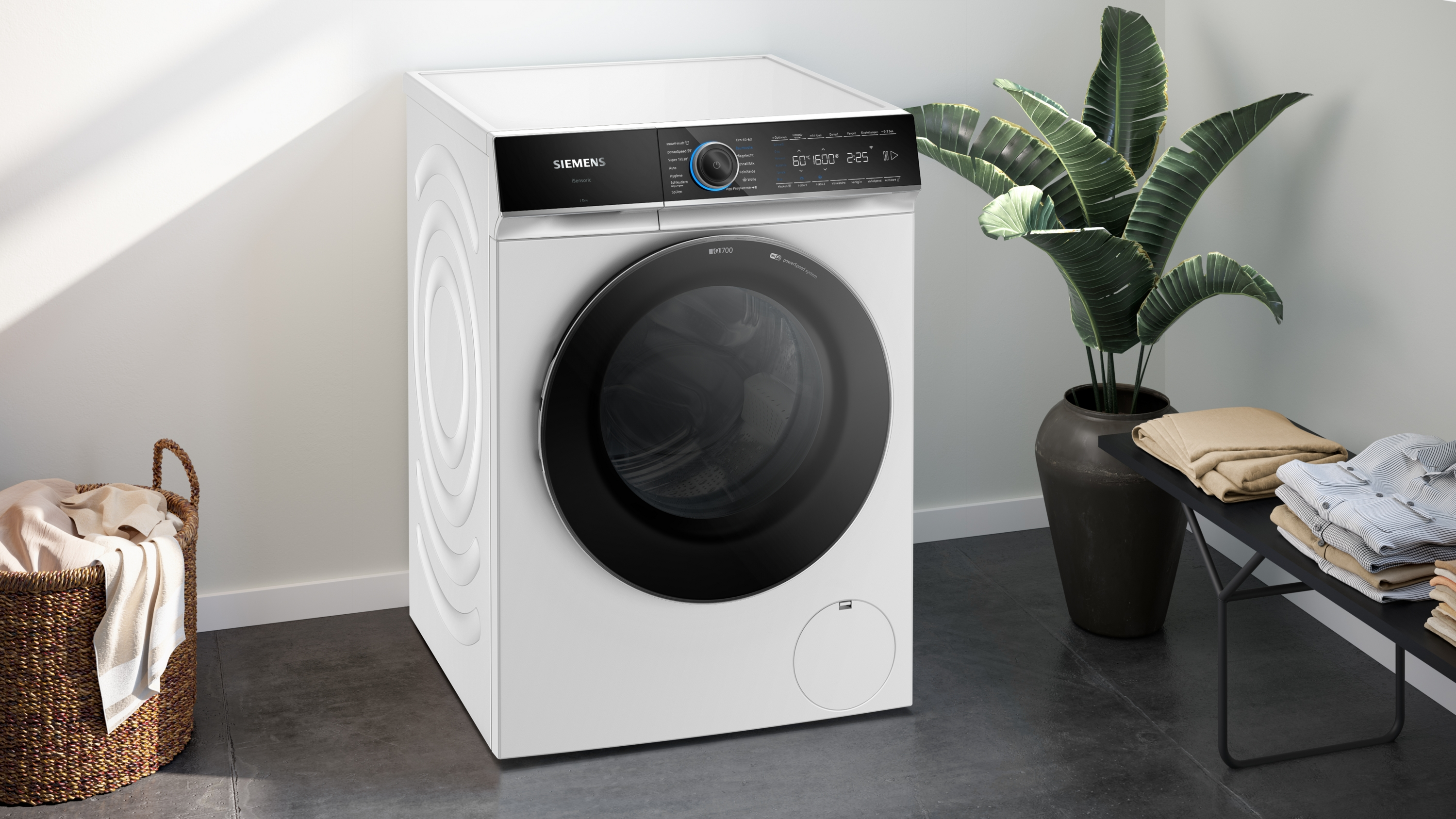 Eine weiße Siemens-Waschmaschine steht in einem Raum neben einer Pflanze im Topf und gefalteter Wäsche auf einer Bank.
