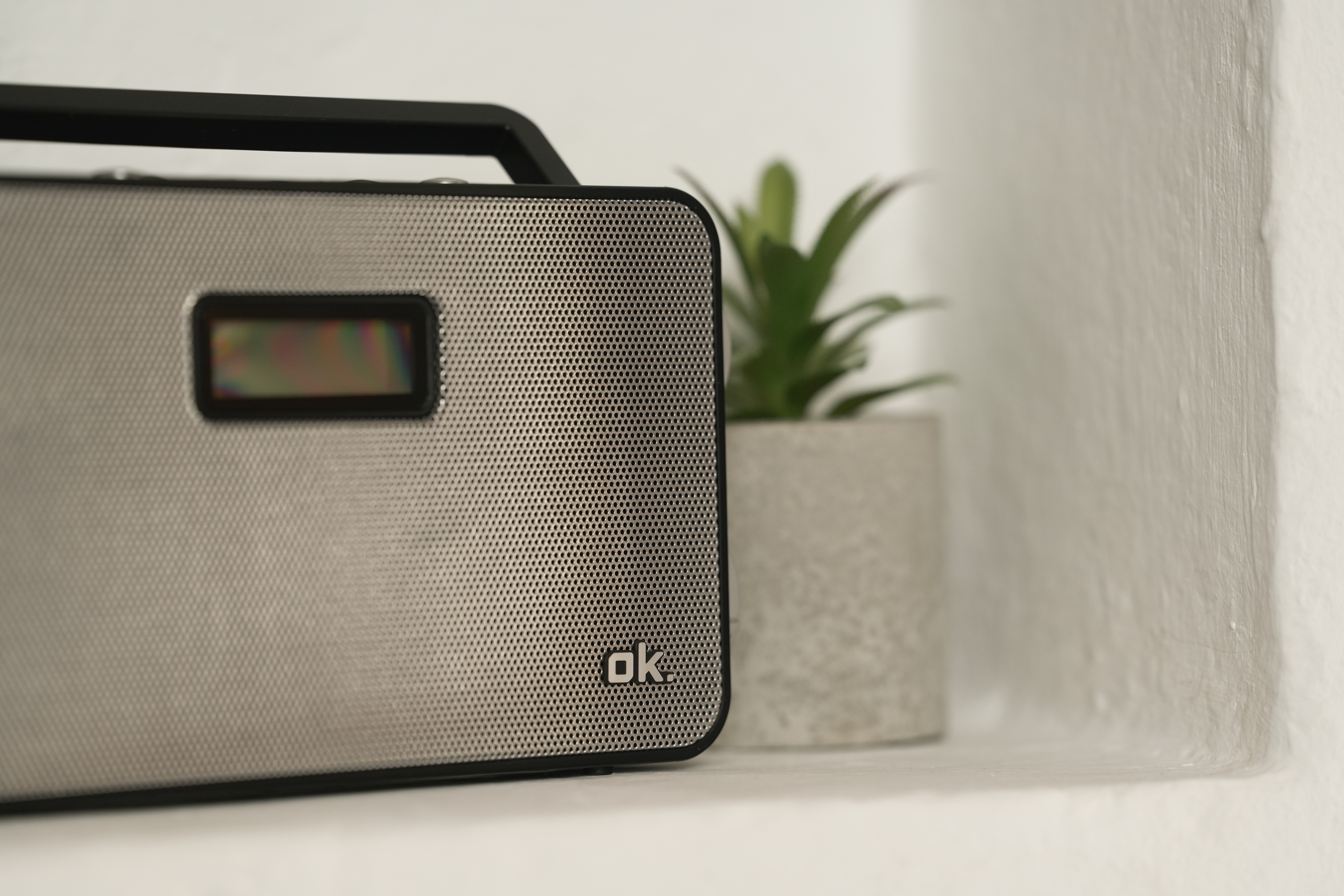 Ein silbernes Radio mit schwarzem Griff und Display steht auf einem Regal neben einer kleinen Zimmerpflanze.