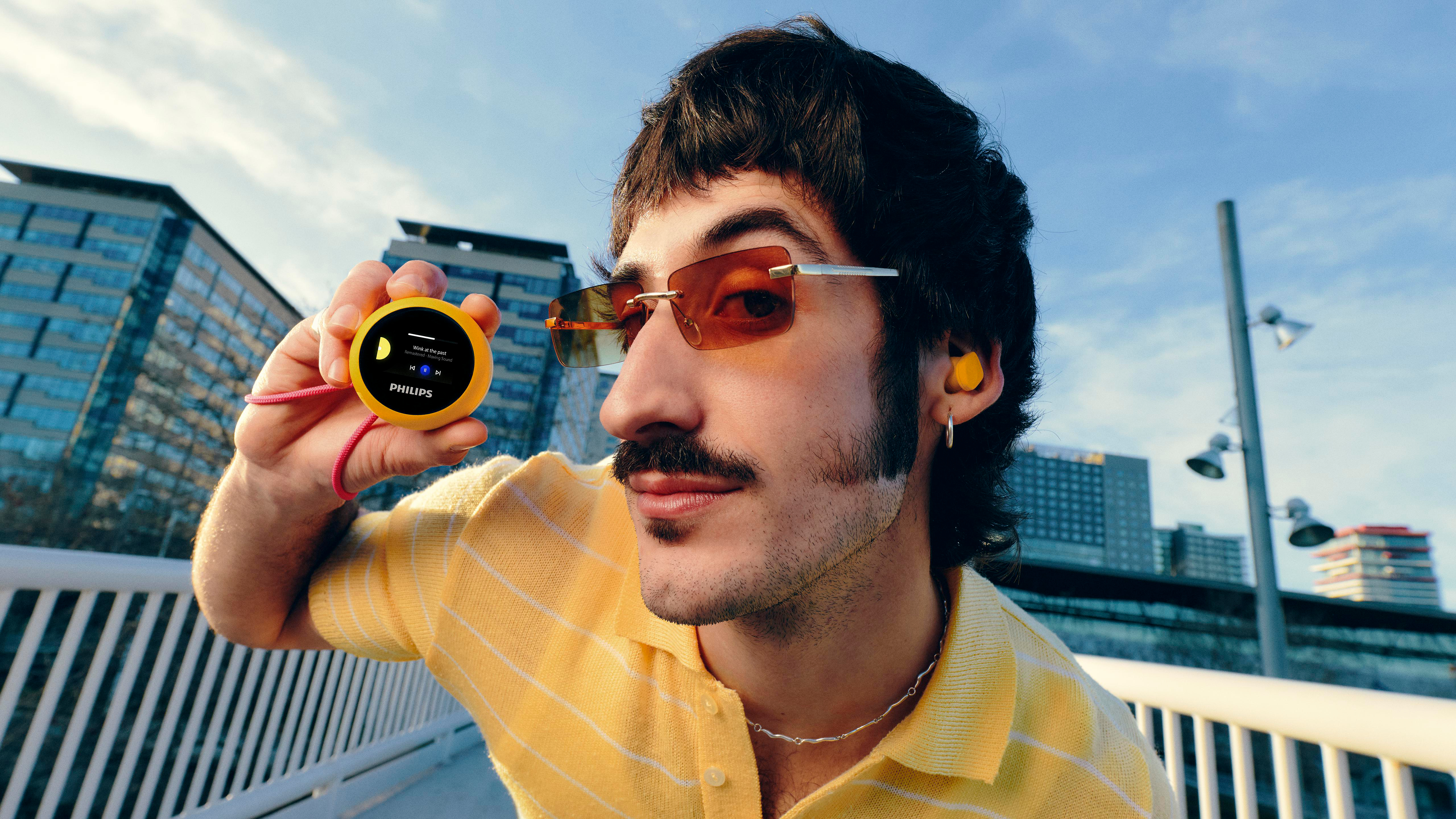 Ein Mann mit gelbem Hemd hält einen gelben Philips-MP3-Player vor einer Stadtkulisse.
