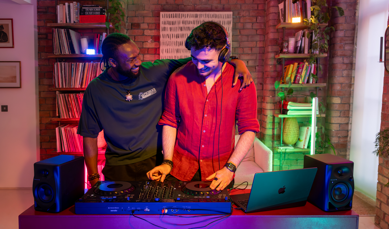 Dos DJ masculinos sonrientes mezclando música en una consola de DJ en una habitación con pared de ladrillo y estantes llenos de discos.