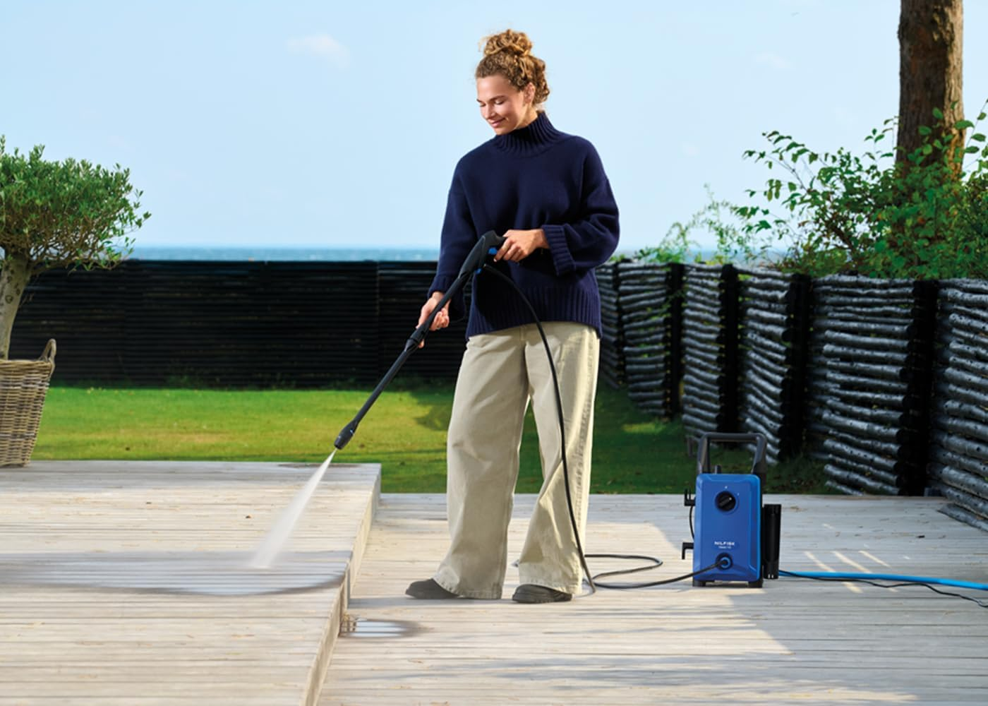 Una mujer con suéter azul y pantalones claros limpia un patio de madera con una hidrolavadora azul.