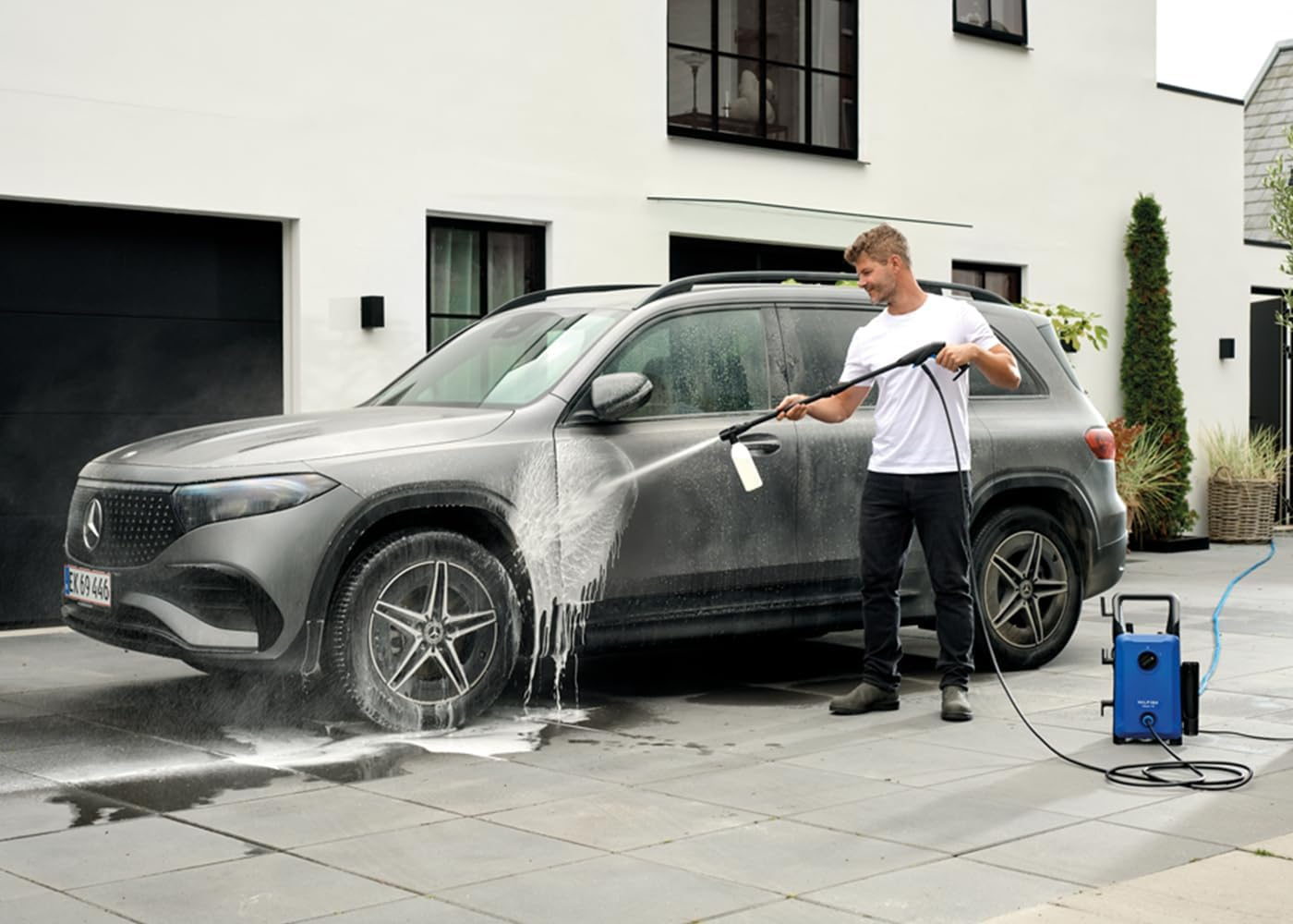 Un hombre lava un SUV gris con una hidrolimpiadora azul frente a una casa blanca moderna.