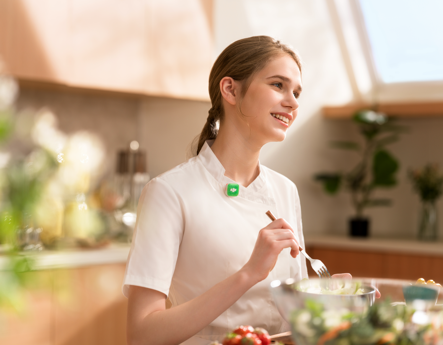 Una mujer joven sonriente con chaqueta blanca de chef come ensalada en una cocina soleada.