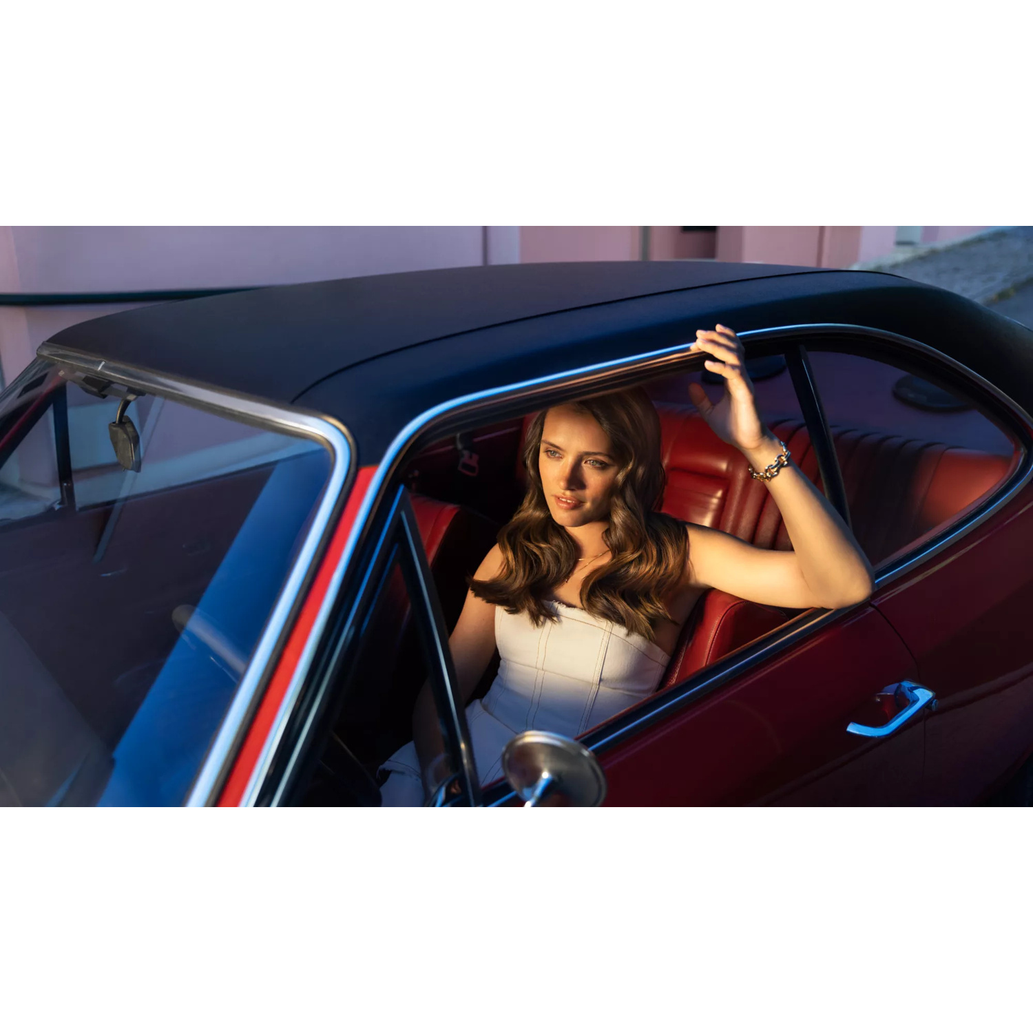 Una donna con top bianco siede in un'auto d'epoca rossa con tetto nero.