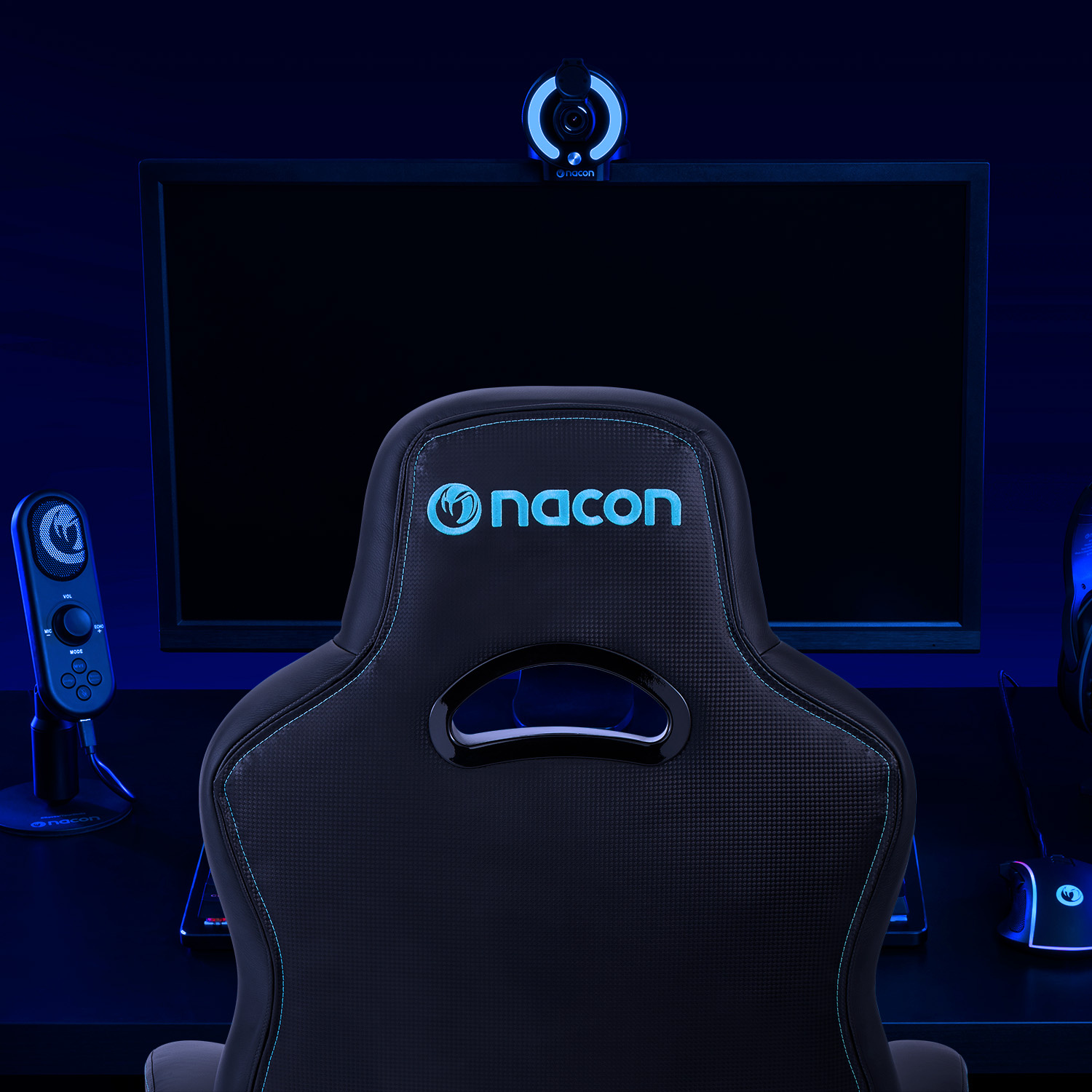 Una sedia da gaming Nacon nera davanti a un monitor, con microfono e webcam in una stanza blu.