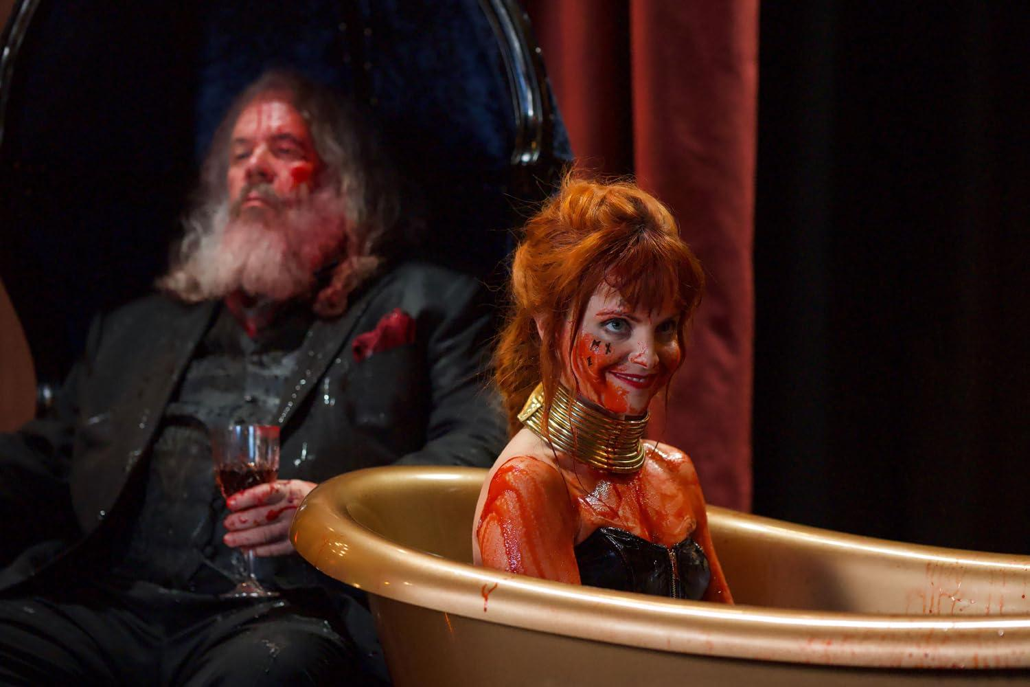 Eine rothaarige Frau mit blutverschmiertem Gesicht und Halsreif sitzt in einer goldenen Wanne, während ein Mann zuschaut.