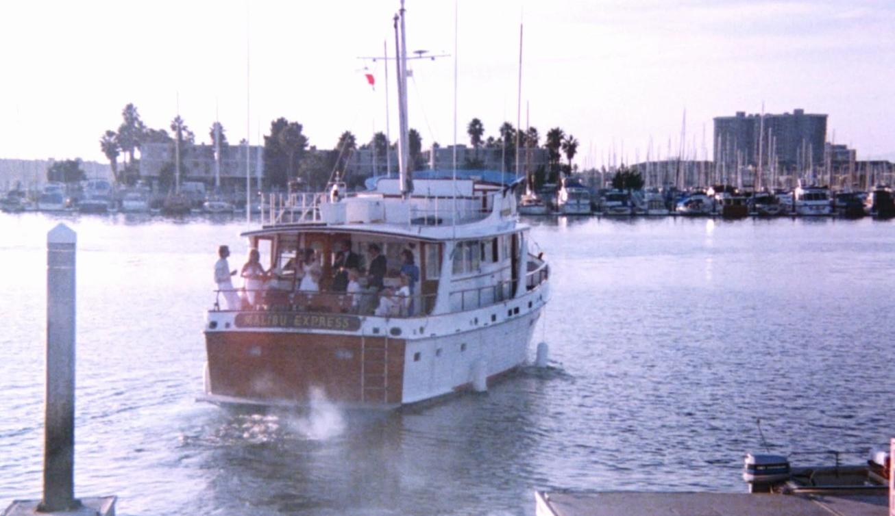 Eine Yacht namens Malibu Express mit Passagieren faehrt in einem Hafen, mit Baeumen und Gebaeuden im Hintergrund.
