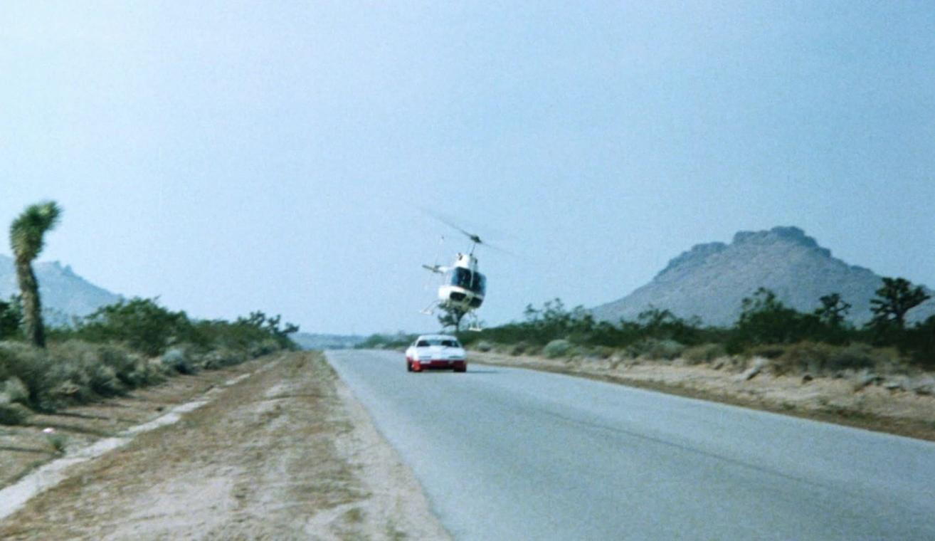 Ein weißer Hubschrauber fliegt niedrig über einem Auto auf einer Landstraße in einer bergigen Wüstenlandschaft.