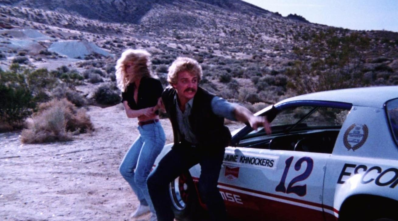 Ein blondes Paar steht in einer hügeligen Wüstenlandschaft vor einem nummerierten Rennwagen.