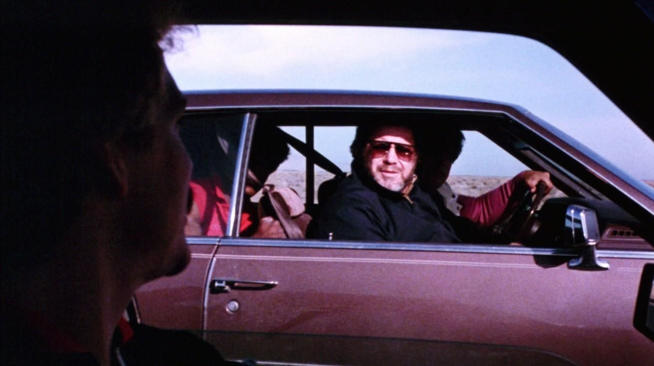 Ein Mann sitzt mit Sonnenbrille am Steuer eines braunen Autos, während ein anderer Mann von vorne zuschaut.