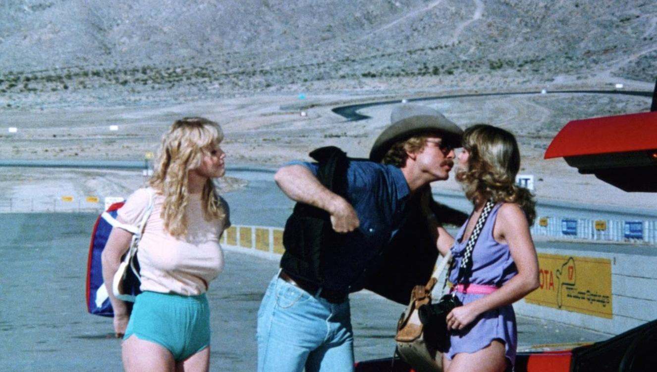 Ein Mann im Cowboyhut küsst eine Frau an einer Rennstrecke, während eine andere Frau zusieht.