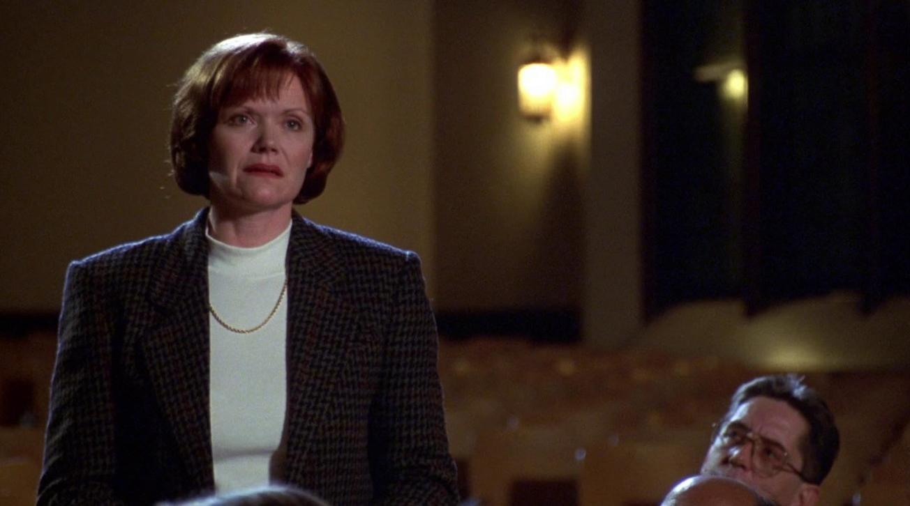 Eine rothaarige Frau mit einer dunklen karierten Jacke steht in einem Auditorium, während andere im Hintergrund sitzen.