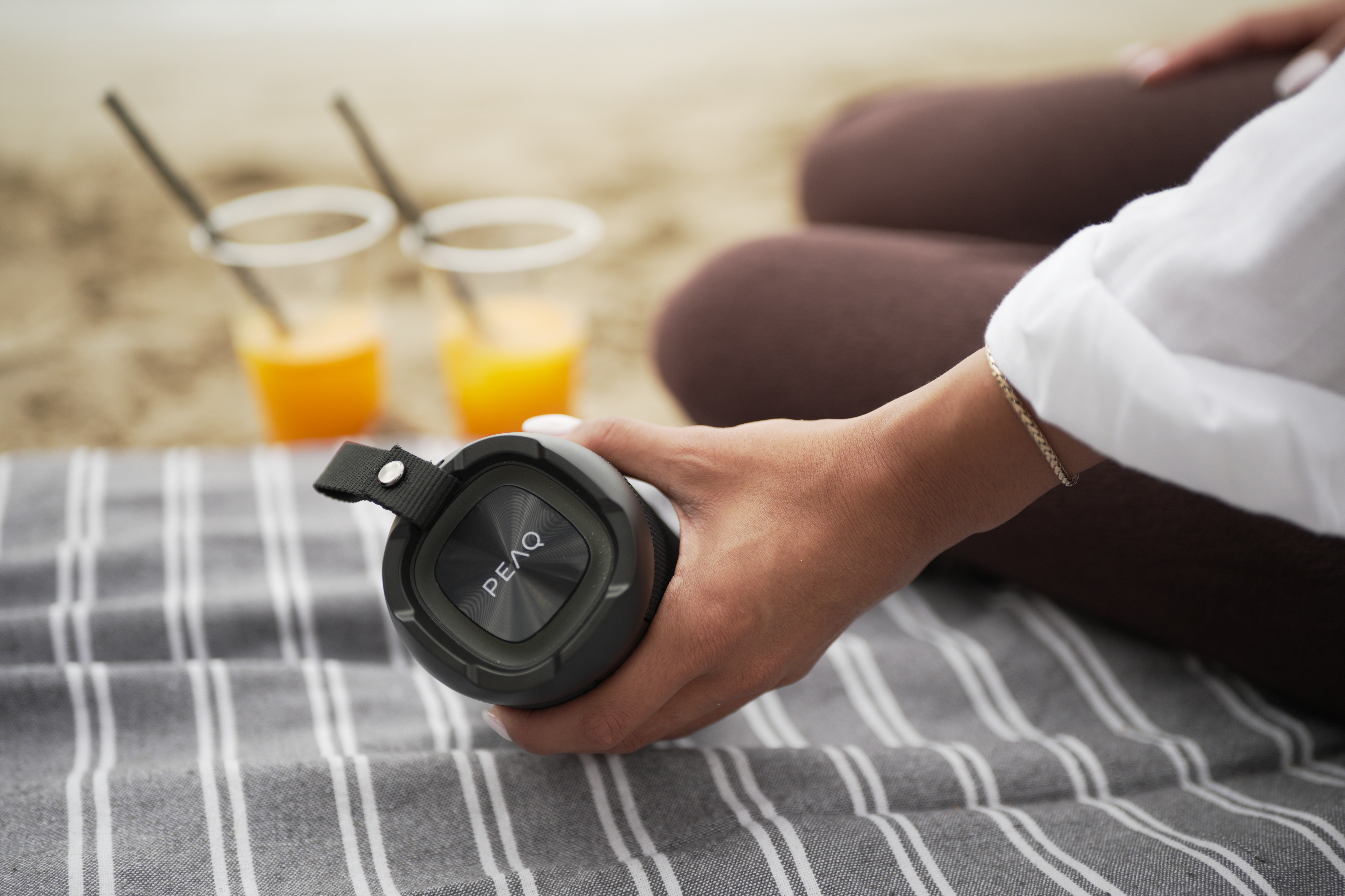 Osoba trzyma głośnik Bluetooth PEAQ na w paski kocu, a w tle na piasku stoją dwa napoje.