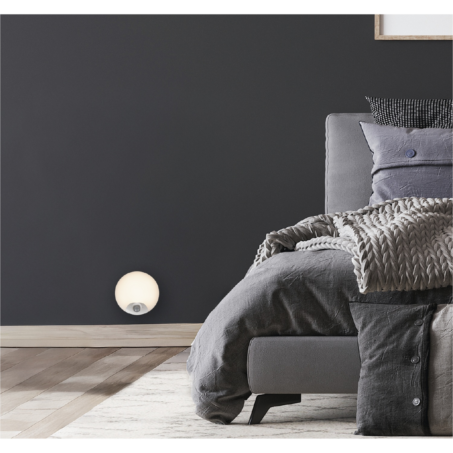Een verlichte ronde wandspot schijnt op een donkere muur naast een modern grijs bed.
