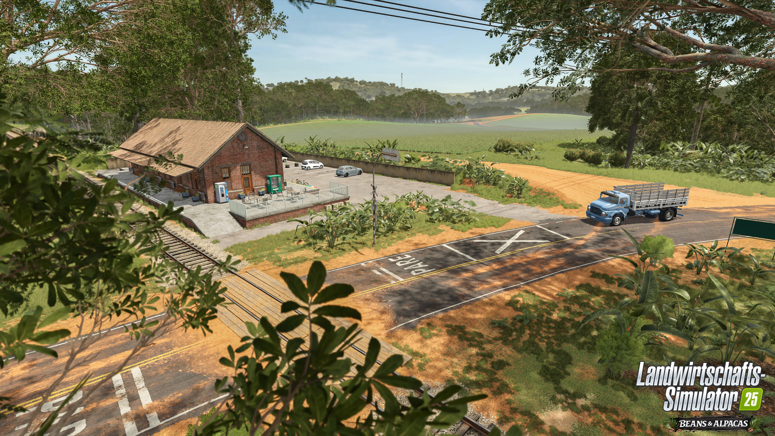 Eine ländliche Bahnhofslandschaft mit roten Backsteingebäuden, Gleisen, einer Straße und einem blauen Lastwagen.