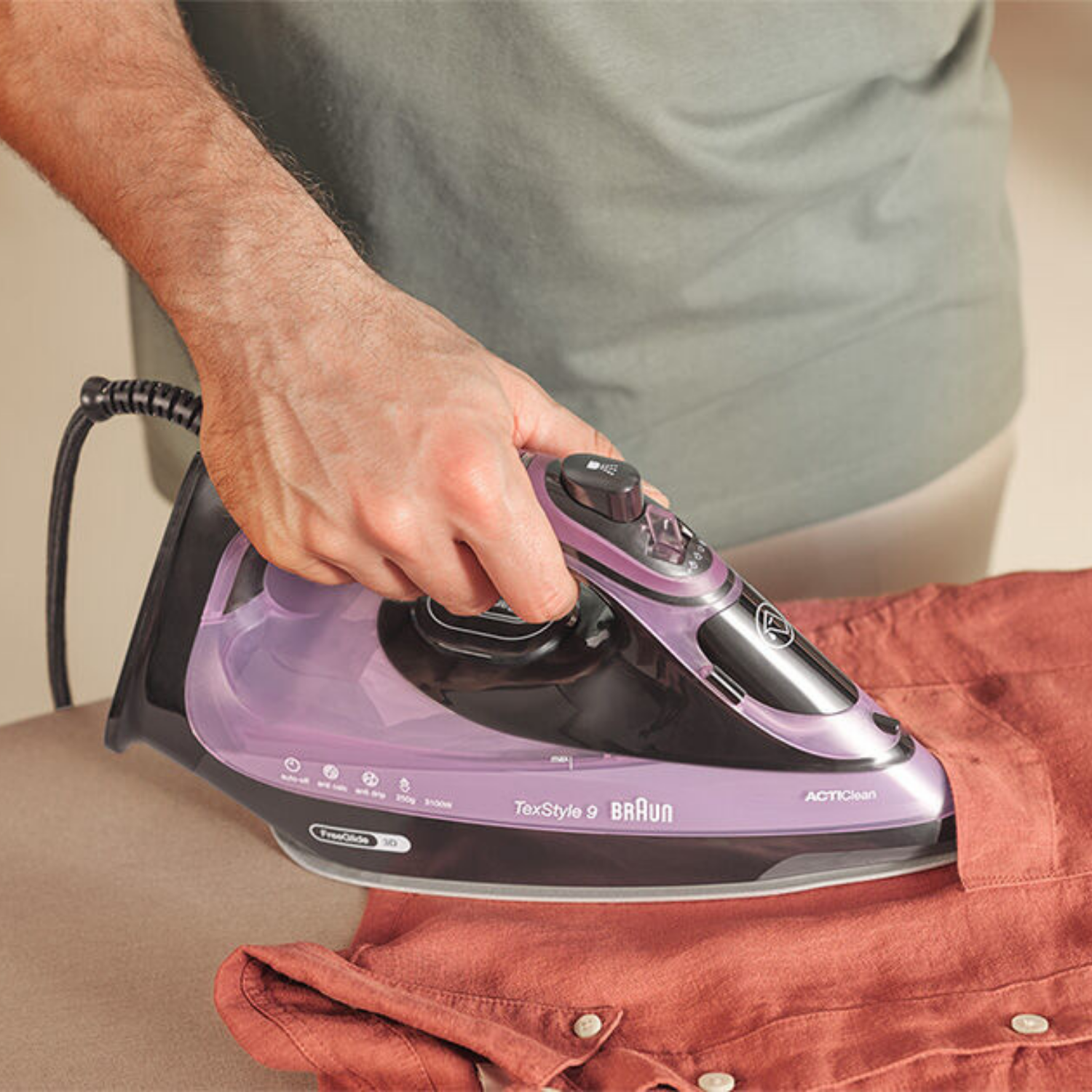 Una persona usa una plancha de vapor Braun morada para planchar una camisa marrón.